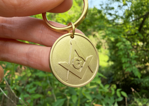 Hand holding a gold keychain medallion featuring the Masonic Square and Compasses symbol with the letter G