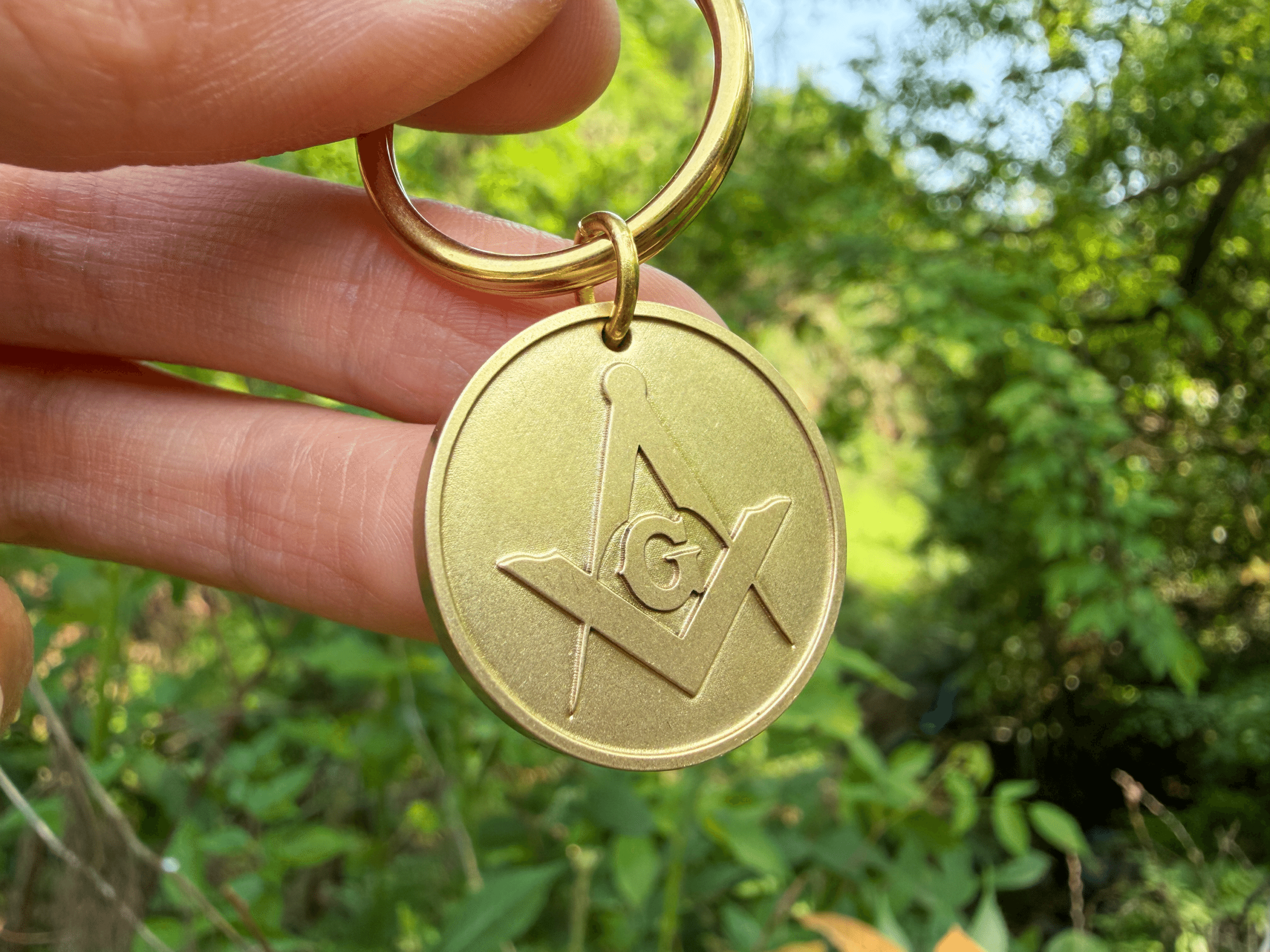 Hand holding a gold keychain medallion featuring the Masonic Square and Compasses symbol with the letter G