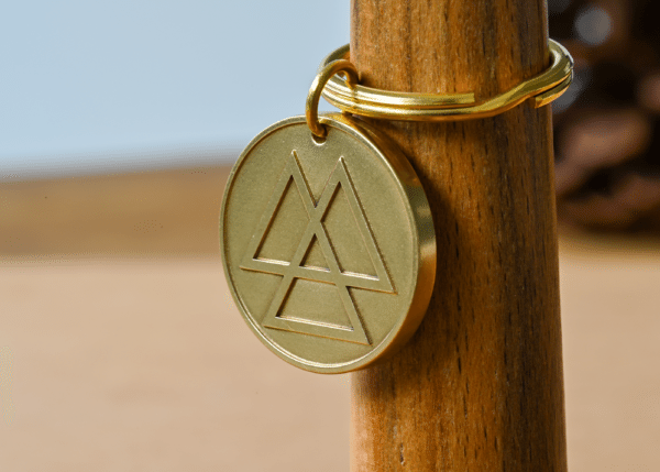Round brass keychain medallion engraved with overlapping triangles hanging from a wooden post