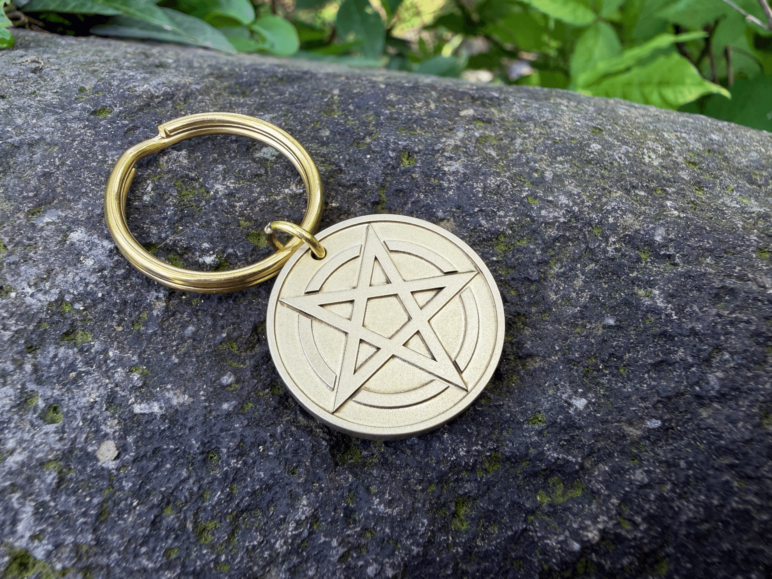Brass keychain featuring a round pendant engraved with a pentagram symbol, resting on a mossy rock