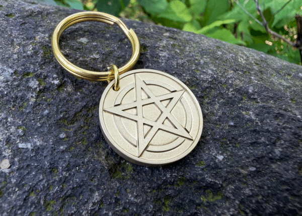 Brass keychain featuring a circular medallion engraved with a pentagram symbol, resting on a dark rock