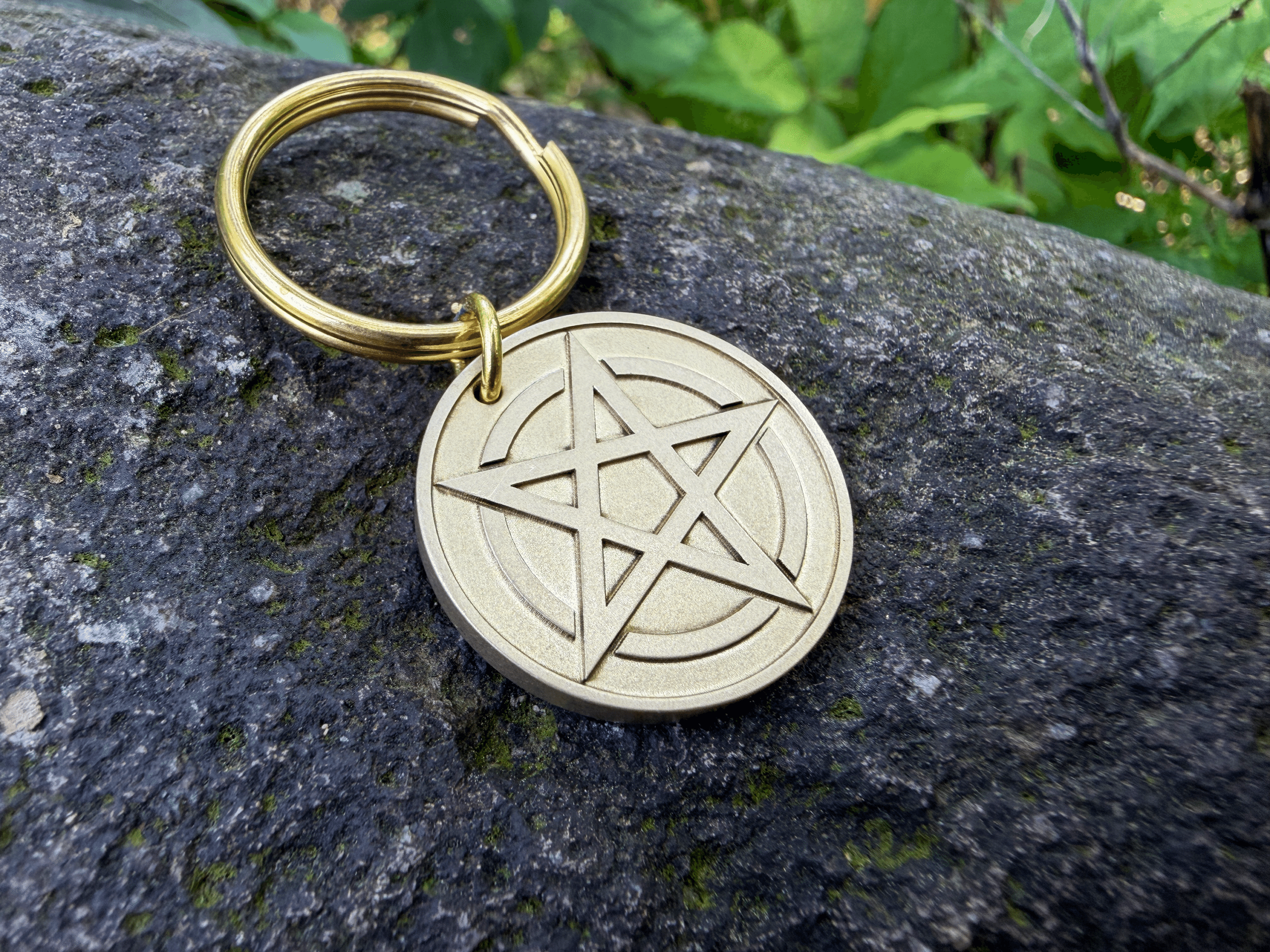 Brass keychain featuring a circular medallion engraved with a pentagram symbol, resting on a dark rock