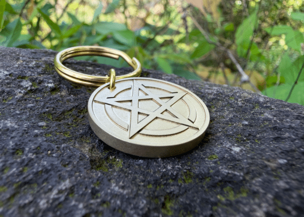 Brass keychain with an engraved pentagram symbol resting on a stone outdoors