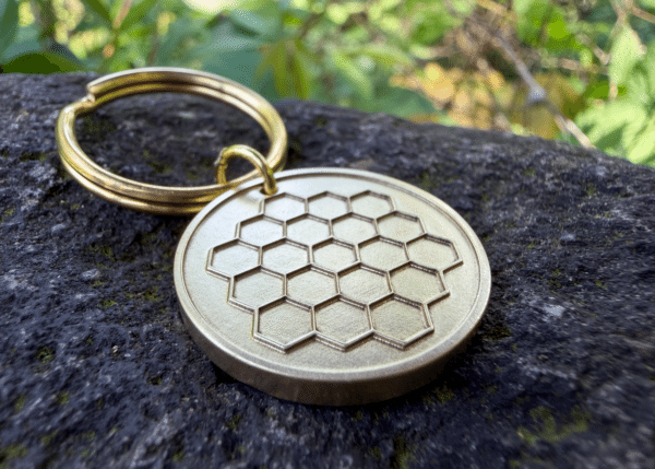 Brass circular keychain tag with an engraved James Webb Space Telescope pattern resting on a stone surface