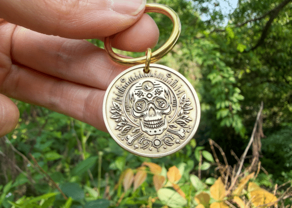 Hand holding a round brass keychain medallion engraved with a sugar skull, crescent moon, and leaves