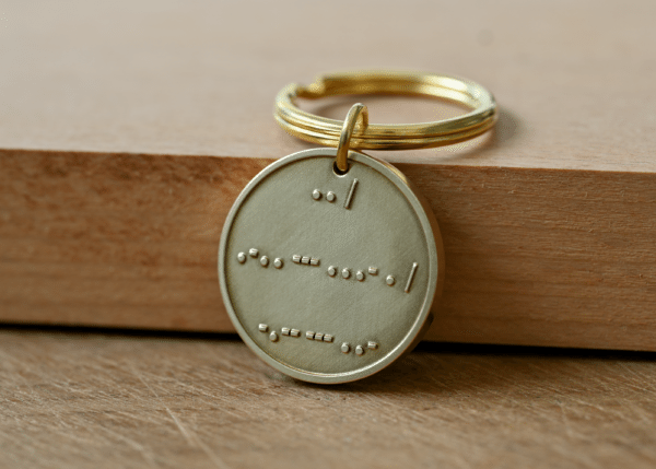 Circular brass keychain tag engraved with Morse code symbols resting on a wooden surface
