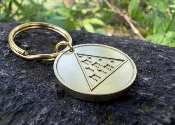 Brass medallion keychain resting on a rock, featuring an engraved triangle with Hebrew lettering
