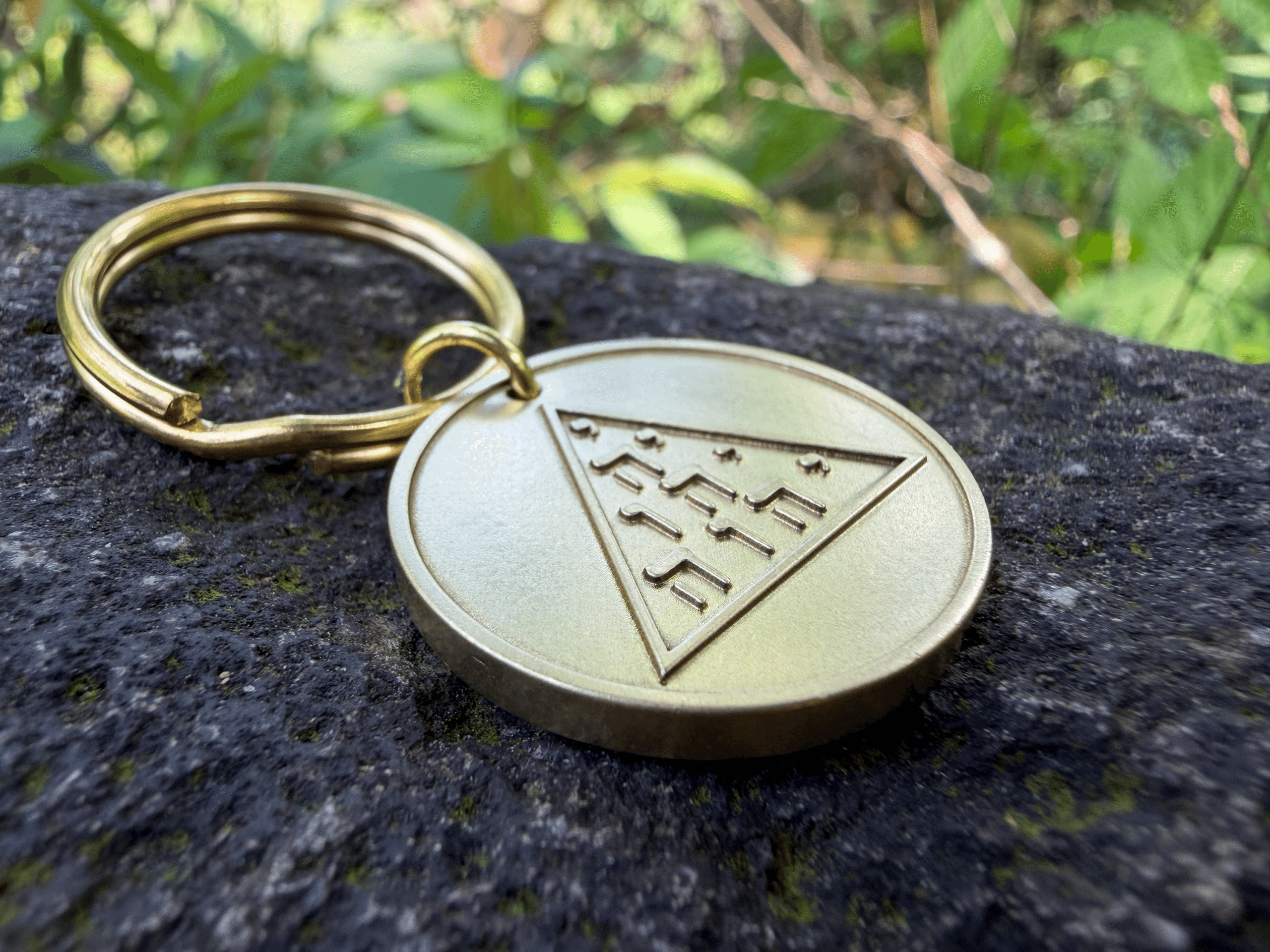 Brass medallion keychain resting on a rock, featuring an engraved triangle with Hebrew lettering