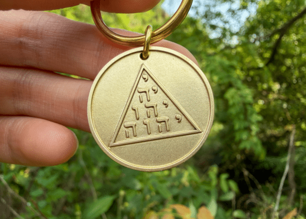 Hand holding a brass keychain medallion featuring a triangle with the Hebrew Tetragrammaton arranged in a tetractys