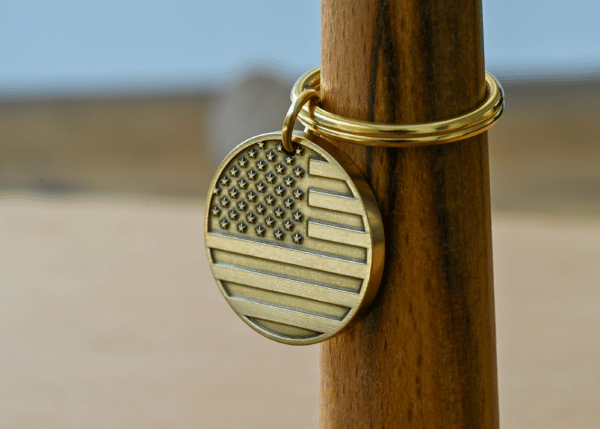 A circular brass keychain tag engraved with an American flag pattern on a brass keyring