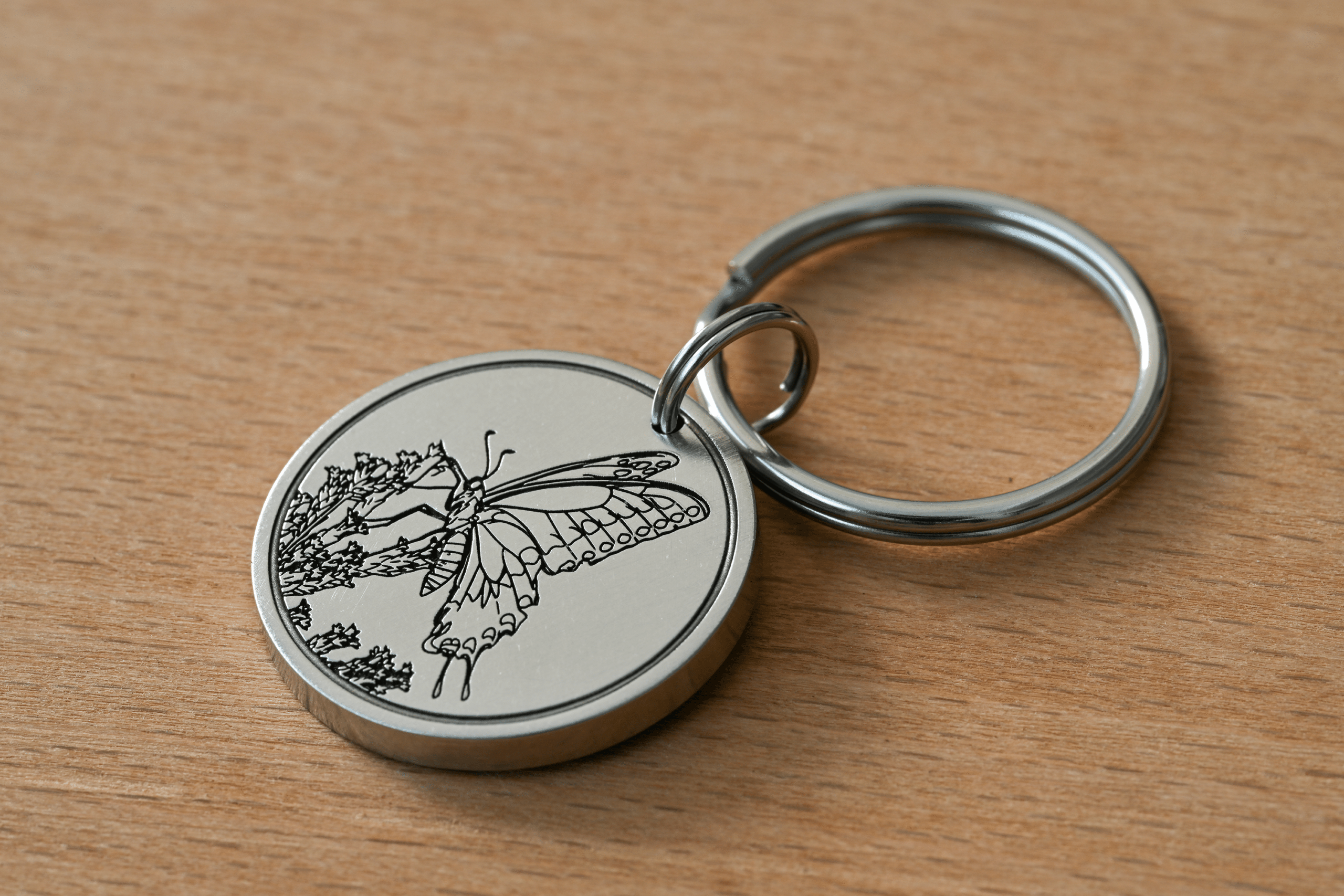 Silver circular keychain engraved with a butterfly resting on flowers, lying on a wooden surface