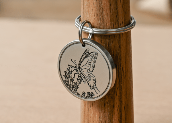 Round silver metal tag engraved with a detailed butterfly resting on flowers