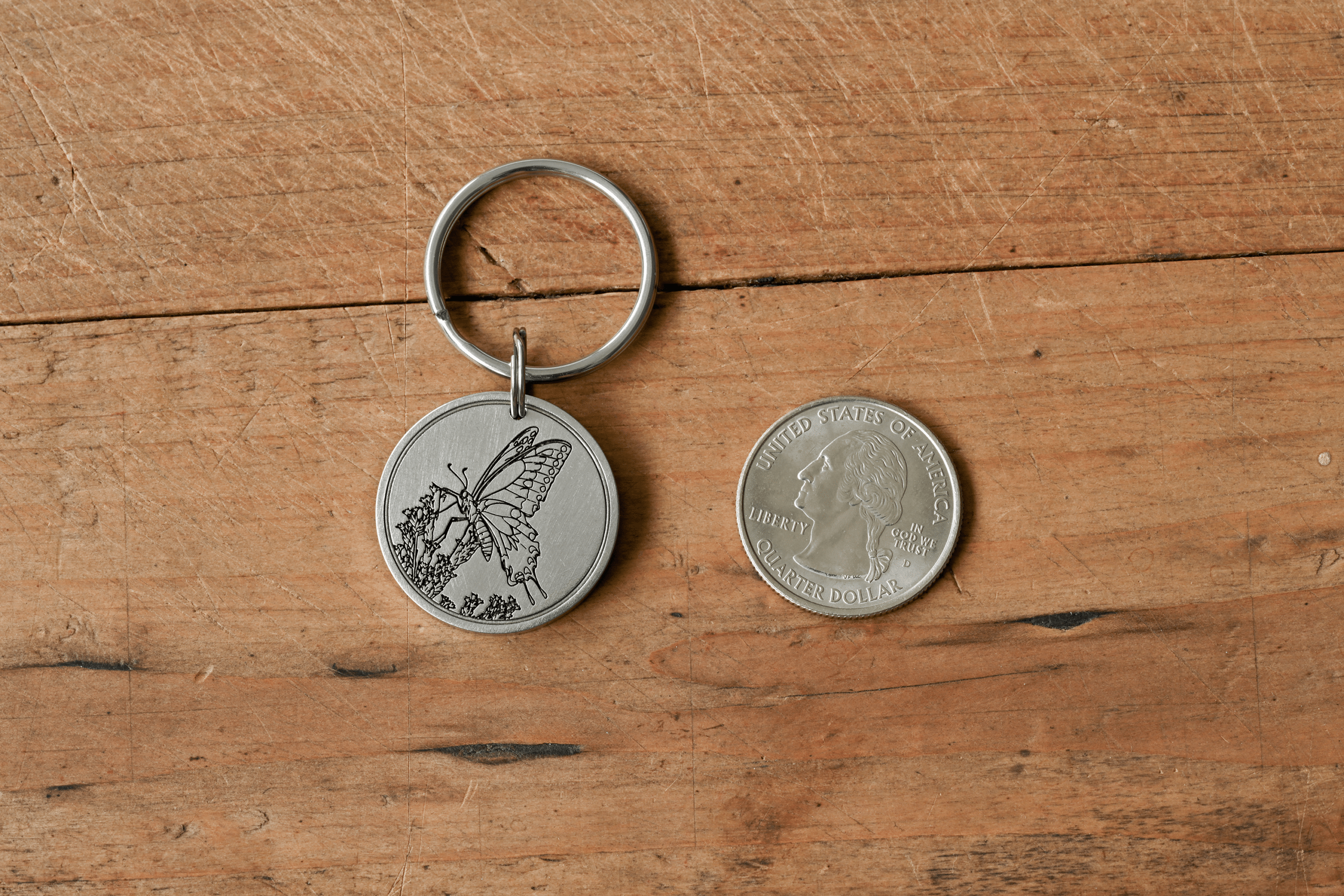 Silver circular keychain engraved with a butterfly, shown next to a US quarter for size comparison